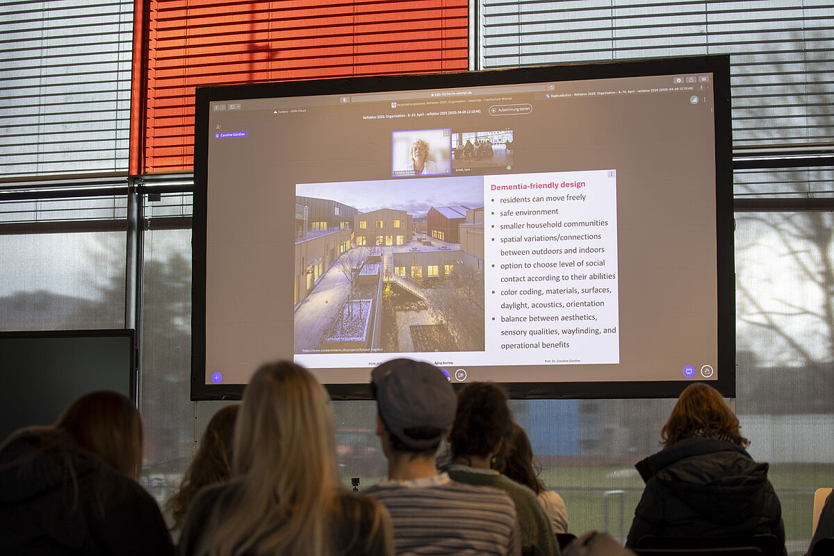 Das Publikum schaut von hinten auf eine Bühne, auf der Caroline Günther per Videokonferenz auf einer großen Leinwand zu sehen ist. Sie hält einen Vortrag zum Thema „Aging Society“.
