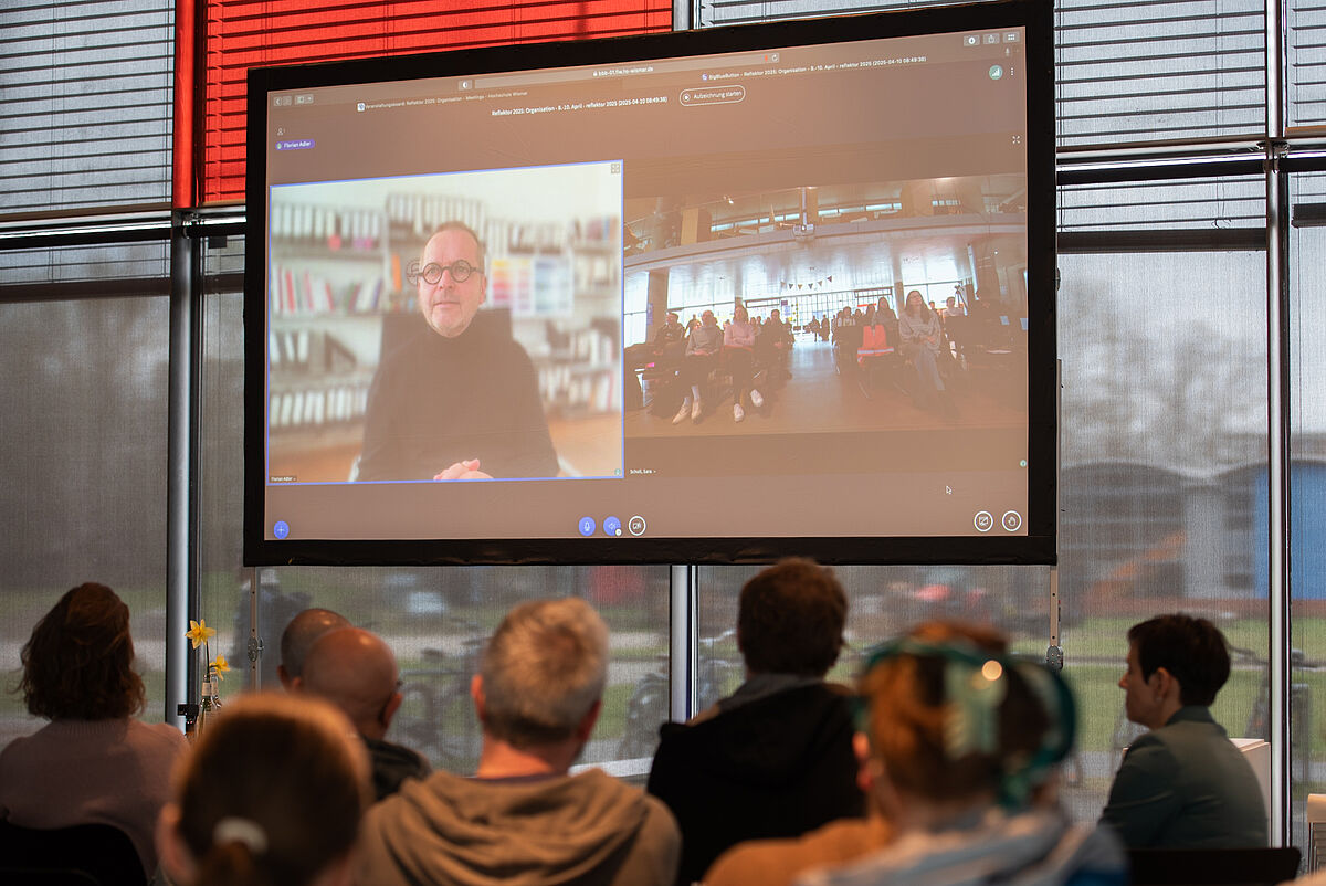 Florian Adler wird digital auf der Leinwand auf der Bühne zugeschaltet, während er einen Vortrag über Inklusives Design hält.