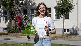 DAAD-Preis 2023 Die Preisträgerin steht auf der Wiese vor einem Hochschulgebäude und hält lachend ihre Urkunde sowie den Blumenstrauß in der Hand.