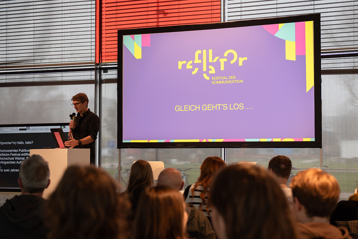 Dekan Oliver Hantke steht auf der Bühne vor Publikum. Rechts neben ihm steht ein großer Monitor, auf dem das Reflektor-Logo und die Worte "Gleich gehts los" abgebildet sind.