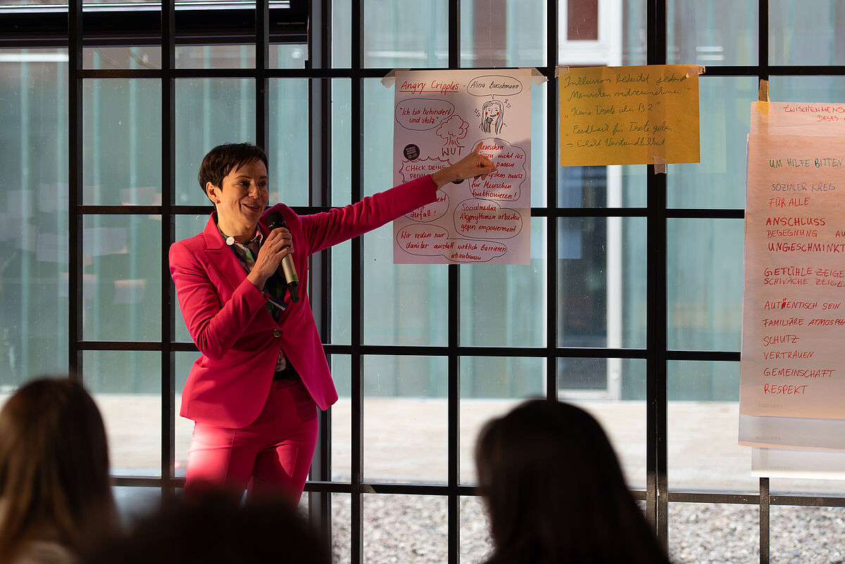 Christine Linke steht vor einem Poster und spricht über die Buchlesung „Angry Cripples“ im Rahmen der Postersession.