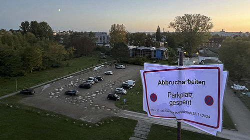Der Sandparkplatz mit seinen vielen Pfützen ist im Abendlicht von oben zu sehen. Im Vordergrund befindet sich ein in das Bild hineinmontiertes Schild mit dem Hinweis auf die Parkplatzsperrung.