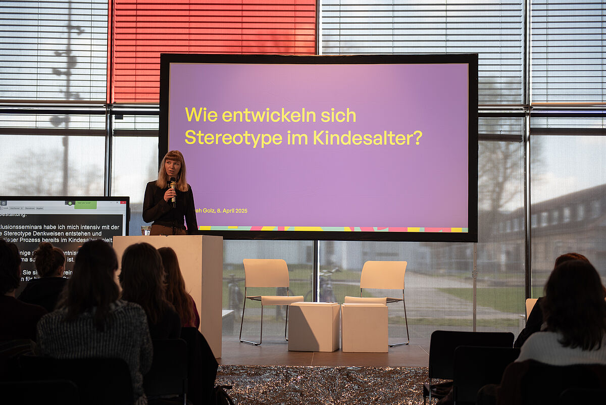 Hannah Golz steht auf der Bühne und hält ihren Vortrag über die Entstehung von Stereotypen im Kindesalter. Das Publikum ist von hinten zu sehen.
