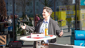 Die Frau steht mit einladen ausgebreiteten Armen hinter einem Tisch. In der Glasfassade hinter ihr sind das Publikum und Teile der leuchtend gelben Ausstellungsstelen zu erkennen.