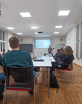 Several people are sitting at tables in a seminar room listening to a lecture. Some are taking notes, others are looking at the screen showing the presentation.