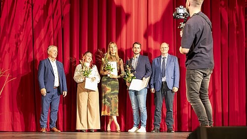 Die Personen stehen auf der Theaterbühne, die rot ausgeleuchtet ist. Am rechten Rand steht ein Kameramann.